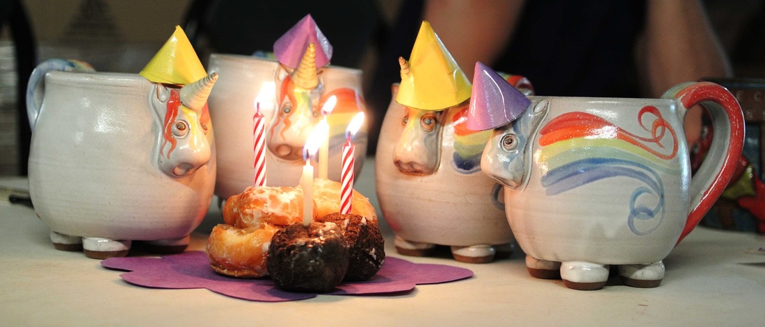 Celebrating Elwood the Rainbow Unicorn turning 4! Four Elwood Mugs wearing birthday party hats are gathered around a small plate of donut holes with four lit birthday candles.